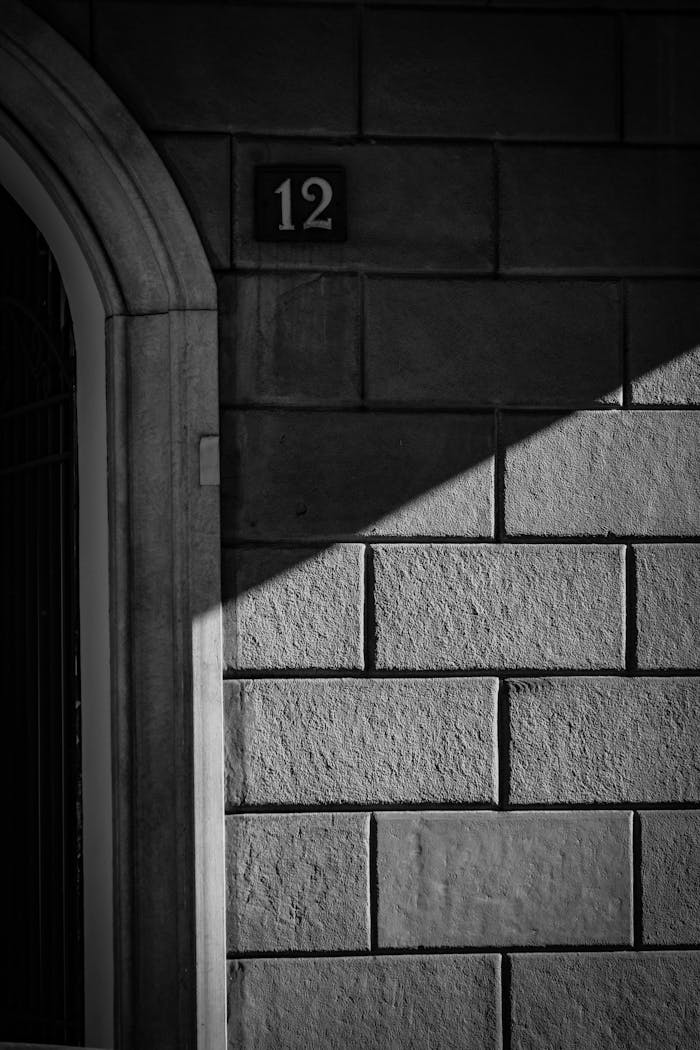 Black and white urban photo featuring a shadow-cast stone wall with the number 12 near a doorway.